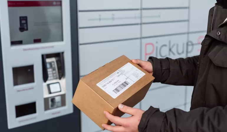 Osoba w ciemnym płaszczu trzyma kartonową paczkę z etykietą obok szarego automatu paczkowego DPD z opcją Pickup.